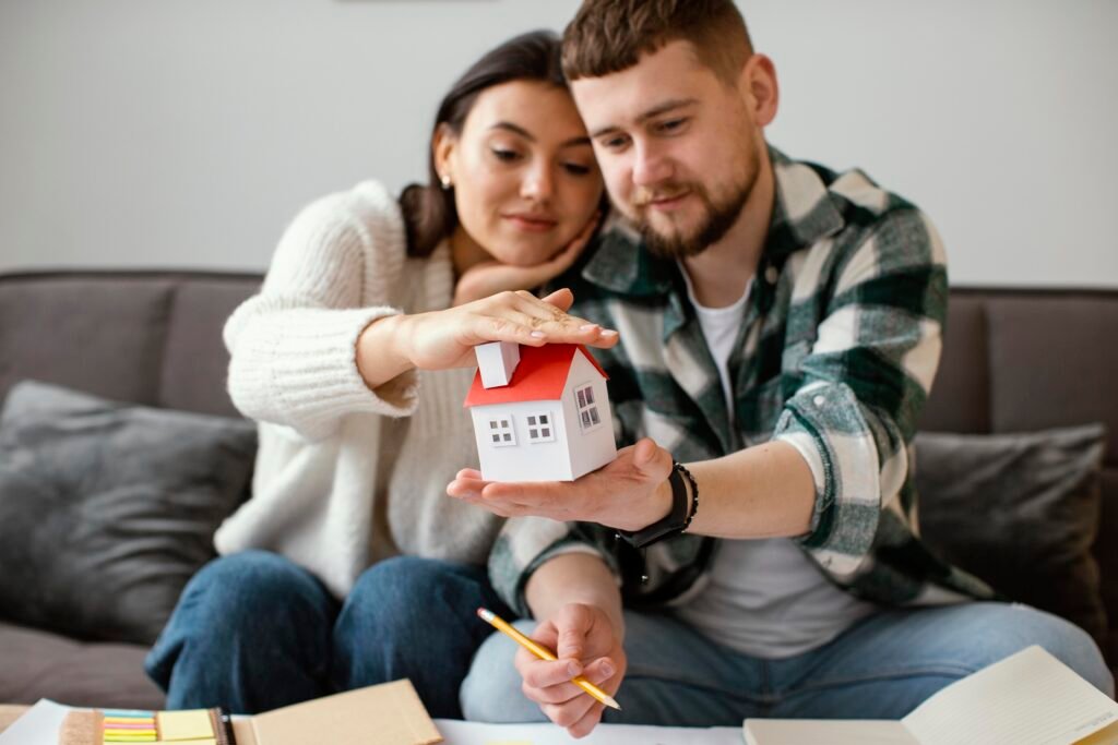A young couple sits closely on a couch, smiling softly as they hold a small paper model of a white house with a red roof between their hands. The woman rests her hand protectively on top of the house while the man supports it from below. Papers, notebooks, and a pencil are visible in the foreground, suggesting they are planning or dreaming about buying a home.