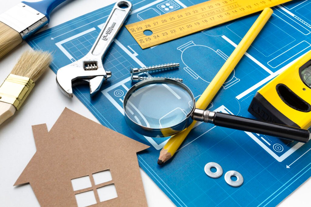 A blue house construction blueprint on a white desk surrounded by essential building tools including a wrench, magnifying glass, yellow ruler, spirit level, and a small cardboard house cutout