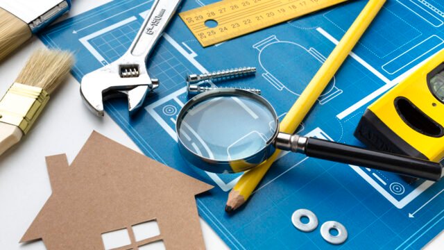 A blue house construction blueprint on a white desk surrounded by essential building tools including a wrench, magnifying glass, yellow ruler, spirit level, and a small cardboard house cutout